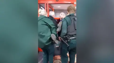 Eileen Mitchell Officers in uniform and a man carry a woman out of a shop. Her legs and feet are being held up by the officers who are carrying her. Shutters are partially down at the doors of the shop.