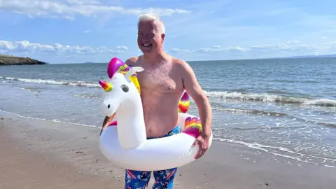 BBC Derek on Barry beach in swimming shorts with a a white unicorn rubber ring around his waist. 