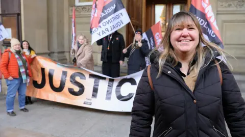 GMB Union A woman in a black puffa coat standing outside a sandstone building with ornate carving on some bricks and stone pillars. Behind her stand four people holding a banner and flags saying "Justice and Fighting for Equal Pay"