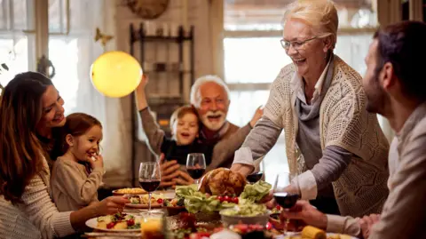 Getty Images Family around the table enjoying a Christmas dinner. Everyone is smiling, as an older woman lays a turkey on the table. 