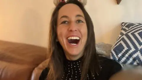 Family photo Amie Gray smiles broadly while sitting on a sofa. She has long, straight, dark brown hair and wears a black top.