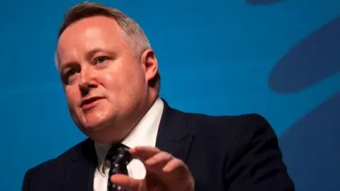 Getty Images Darren Millar is stood in front of a blue background, appearing to give a speech. He is photographed at an angle and is wearing a black suit, a tie and a white shirt.