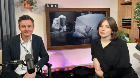 BBC Andy Howard and Marie Lennon sat in their podcast recording studio. Andy has dark brown hair and is wearing a white shirt and blue checked blazer. Marie has a mid brown bob haircut and is wearing a black dress. 