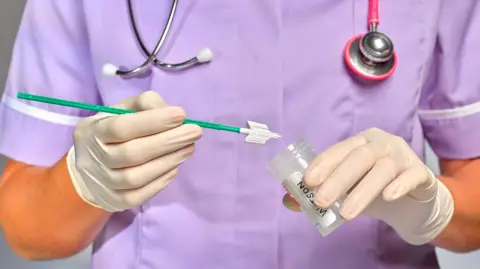 Getty Images Image of a nurse in purple scrubs, holding smear test appliances, a test tube and wearing white gloves. They have a stethoscope around their neck.