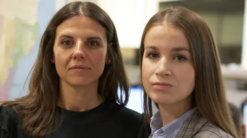 BBC Spanish elite policewomen Cristina and Lidia look at the camera. Cristina is wearing a black top and has long brown hair. Lidia also has long brown hair and is wearing a blue shirt and checked jacket.