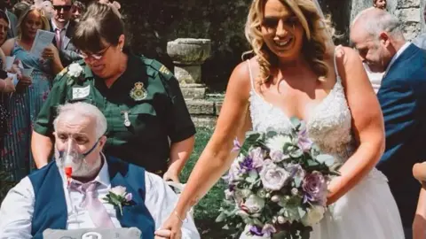 CJ Photography Wales Wayne is in a wheelchair, wearing an oxygen mask. He is being pushed down the "aisle" of the outdoor wedding by a paramedic in uniform. His daughter is dressed in a wedding dress with bouquet and is holding his hand.