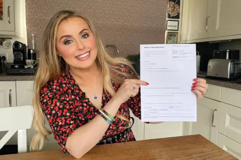 BBC Rosey Hudson holds up her notice of discontinuance while sitting at her kitchen table, she's smiling and wearing a floral red top with red painted fingernails