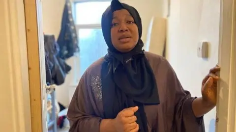 BBC Fauzia is standing in a doorway in her house. She is wearing a black headscarf covering her head which is draped around her neck. She is wearing brown clothing. Behind her you can see a bedroom.