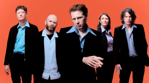 Carry On PR The band Franz Ferdinand (four men and one woman) are looking towards the camera. All are dressed smart casual, with a bright orange background behind them.