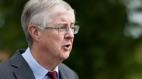 Getty Images Welsh government minister Mark Drakeford wearing glasses and a red tie, speaking in front of a blurred green background