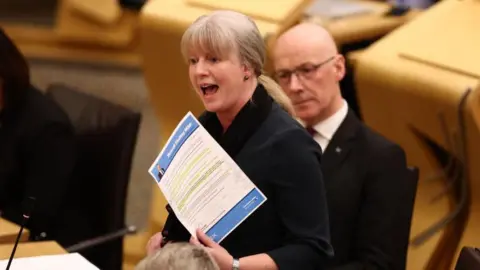 Getty Images Shona Robison holds a piece of paper up in the Holyrood chamber. Her blonde hair is back in a ponytail and she wears a black blazer. John Swinney can be seen behind her - he wears a white shirt, dark red tie and a black suit jacket.