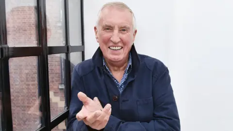 BBC Johnny Walker smiling at the camera in BBC Old Broadcasting House. He is wearing a blue and white checked shirt and navy jacket.