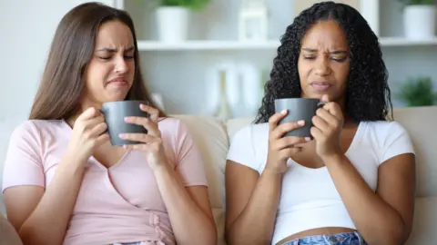 Getty Images Two women looking distastefully in to their mugs whilst sat on the sofa