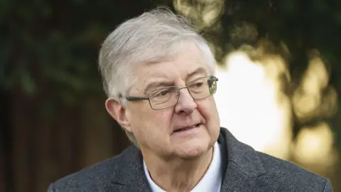 PA Media Mark Drakeford is wearing a grey suit, white shirt and glasses in front of a blurred background