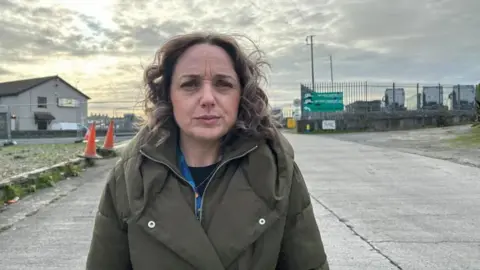 BBC/Carwyn Jones Llinos Medi looks concerned as she stands on a side road by Holyhead port, with some HGVs parked up in the background. She is wearing a camo green coat and a black top. She has curly blonde shoulder length hair.