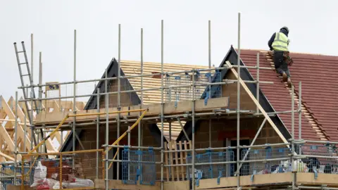 PA Media A construction site showing a house in progress, with scaffolding surrounding the structure and a worker in a high-visibility vest installing roof tiles on the sloped roof. Wooden beams and other construction materials are visible in the foreground and background.