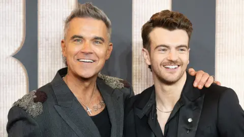 Getty Images Robbie Williams has a huge smile and his arm around Jonno Davies who is also smiling. Both smartly dressed at a premiere event for Better Man.