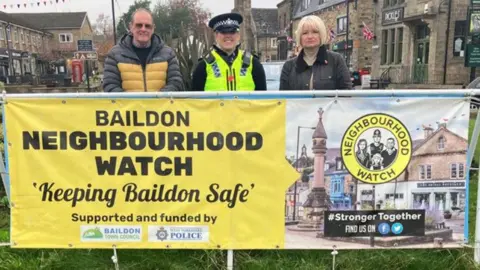 BBC A police officer and two members of the public stand behind a sign on a grassed roundabout, which reads 'Baildon Neighbourhood Watch: Keeping Baildon Safe'.