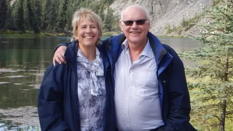 Liz Barlow Mrs Barlow and Mr Greenaway stand in front of a lake, surrounded by pine trees. The couple smiles at the camera. Mr Greenaway has his arm around his wife's shoulder.