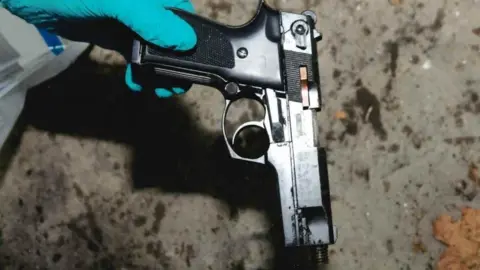 Crown Office Close up of a forensic officer, wearing a white hazmat suit and blue latex gloves, holding a black pistol by the handle with the barrel of the weapon pointing at the ground. 