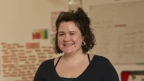 Catrin-Mai Huw smiles at the camera, dressed in a black top, with short curly brown hair, stood in a classroom