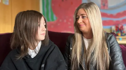 BBC Hannah and Ashly are sitting in a classroom looking at each other. Ashly, the mother, has long blonde hair and is wearing a grey leather jacket and white blouse. She is smiling at her daughter who is looking into the distance. Hannah has shoulder-length dark hair and is wearing a white school shirt without a tie and black hoodie. 