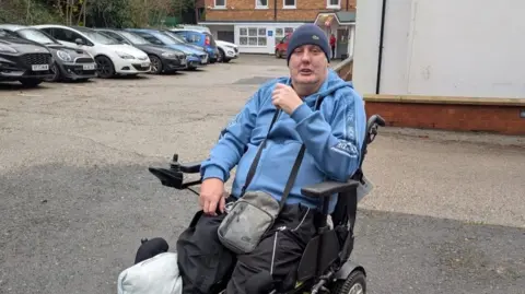 Chris Smith Chris is in a car park in his electric wheelchair. He is wearing a light blue hoody and hat