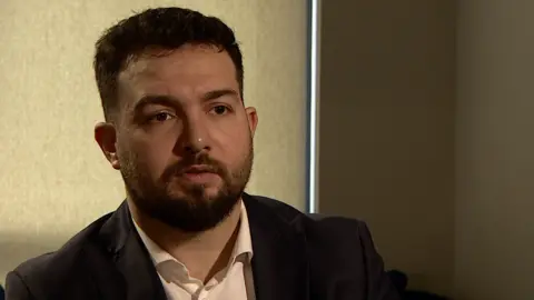 BBC A man in a white shirt and black suit jacket sitting in a darkened room with the blind drawn. He has dark short hair and a dark beard.