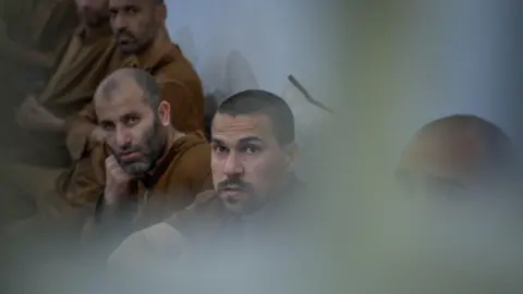 BBC/Matthew Goddard Six male inmates sit together in a row against a wall, looking in the same direction, all dressed in the same brown clothing