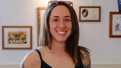 Craig Blake Amie Gray stands in a room with pictures on the wall. She has long, straight dark brown hair. Her black vest top shows tattoos running down both arms.