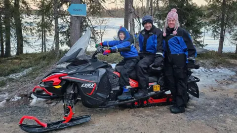 Lisa O'Neil Wayne O'Neil and his son Finn sitting on a snowmobile with Lisa O'Neil standing next to them. They are all wearing snow suits and woolly hats, but there is no snow on the ground around them. There is a lake in the background.