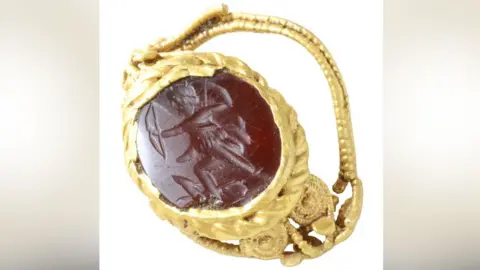 Andrew Williams/Norfolk County Council The front of a gold Roman ring, with a red stone carved with a view of Diana pulling back her bow set in its middle, surrounded by its gold setting. The gold of its crumpled setting and band is ornately worked 