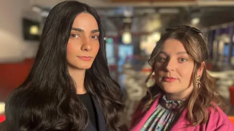 BBC Ellie Wilson & Hannah McLaughlan are photographed looking directly at the camera. The background is blurred out but it is the BBC Scotland newsroom.    Ellie has long dark hair and is wearing a dark suit. Ellie is smaller, with shoulder-length hair and a side-parting. She is wearing a pink jacket and has a colourful shirt. 