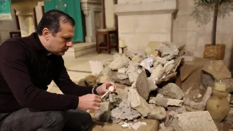 Reuters Reverend Munther Isaac lights a candle near a pile of rubble with baby Jesus figure lying on top inside the Evangelical Lutheran Church in Bethleham