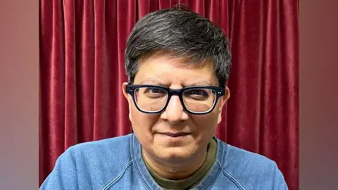 Anthony Abdool Anthony Abdool wearing glasses, a blue sweater over a green T-shirt, in front of a dark red curtain