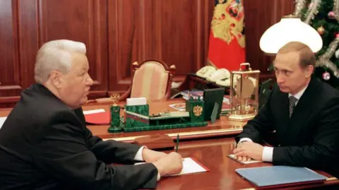 Getty Images Boris Yeltsin sits opposite Vladimir Putin at a table, looking at each other, the day Yeltsin annouced he was stepping down as president