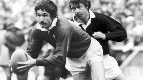 Getty Images Gerald Davies on Murrayfield rugby pitch in 1975. The picture is in black and white and shows Davies running while carrying a rugby ball, and another player close behind him