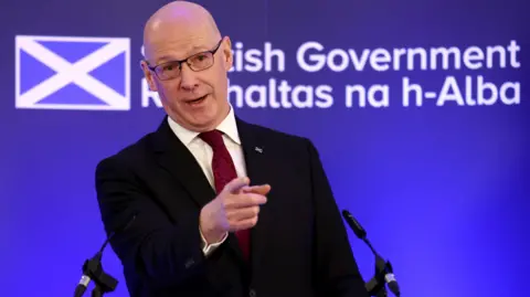 PA Media John Swinney points with his right hand while standing at a podium. He is wearing a black suit, cream shirt and a burgundy tie. There is a blue background featuring a Saltire and the Scottish goverment's logo.