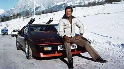 Getty Images Sir Roger is sitting on the bonnet of a black and red Lotus Esprit Turbo sports car. Behind him are snow covered mountains. Sir Roger is wearing a warm white puffy jacket, brown trousers and brown gloves. The numbers "007" can be seen written on his jacket. On the back of the car, are two sets of skis.