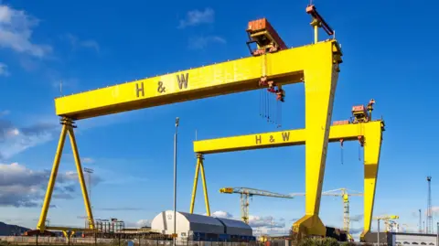 Getty Images Two large yellow cranes with H & W written on them. Below them some smaller cranes and warehouses. Above them blue skies with a few clouds.