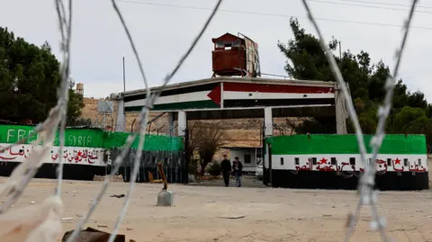 Reuters View of a prison through barbed wire, with two people walking and walls painted with 'Free Syria' and the flag colours