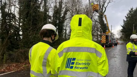 BBC NIE workers in hi-viz hello jackets watching a cherry picker repair a damaged electric pole