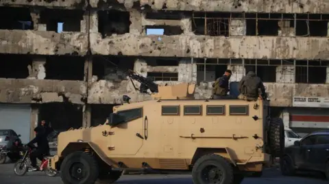 EPA sand-coloured armoured car in front of a ruined building in Homs