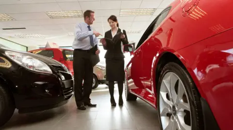 Getty Images Man and a woman in smart clothing talk in a car dealership, standing between new cars