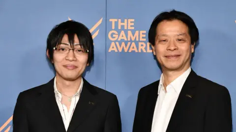 PictureGroup Two men in black suits and white, open-collared shirts, pose for a photo in front of a Game Awards hoarding. The younger man on the left has a mop of black hair and wears glasses, while the older man on the right has a shorter, neatly trimmed style.