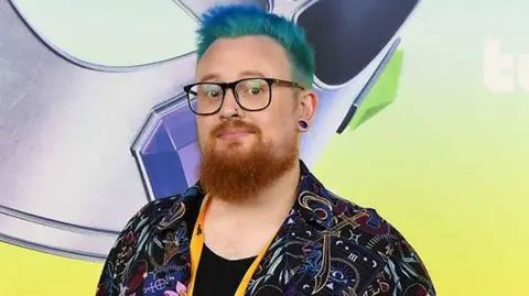 Warwick A person looks at the camera with dyed blue hair and a ginger beard. They're wearing glasses and have a nose piercing