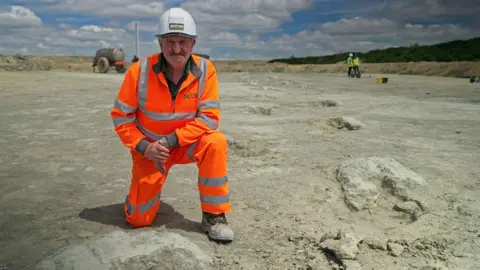 BBC/Kevin Church Gary Johnson a man looking to be in his sixties with a determined state and a grey moustache, dressed in an orange jumpsuit and sand covered boots with a white helmet, kneels with one knee up, one knee on the ground next to the dinosaur footprints he found. They are large craters of indistinct shape in this picture, which trail off into the distance in the whitish-grey sand of a quarry. In the distance behind him to the right two people in yellow hi-visibility waistcoats and hard hats stand with buckets beside them on the ground.