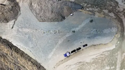 BBC/Kevin Church Qn overhead drone shot taken from about 200 metres up shows a large quarry with the two sets of dinosaur prints criss-crossing it. There are also several vehicles, a couple of tents and about 15 workers in yellow hi-visibility clothes.