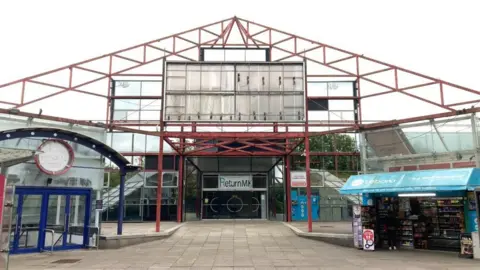 The Point in Milton Keynes. A large red metal pyramid structure with grey paving in front of it. The site is abandoned but an open kiosk is to the right at the front