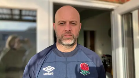 James Richardson stands outside his home, looking at the camera. He is wearing a navy blue T-shirt. He is balding and has a grey-brown beard and blue eyes.